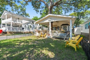 A covered outdoor seating area with chairs and a table, a white building with a porch, and a large tree.
