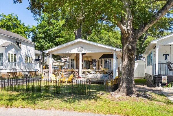 A mobile home with a porch, a fenced yard, and a large tree.