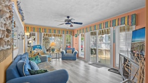 Ein geräumiges Wohnzimmer mit einem blauen Sofa, einem Sessel mit Strandkissen, einem Deckenventilator und einem großen Fenster mit Jalousien.