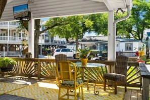 A covered outdoor seating area with wicker chairs and a glass table, overlooking a street with buildings and trees.
