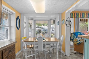 A dining area with a wooden table and white chairs, a bay window with blinds, and a colorful curtain.