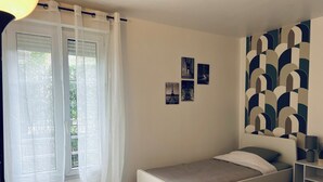 A bedroom with a single bed, a window with sheer curtains, and a wall with a geometric pattern. - Appartement Spacieux et Calme (Bussy-Saint-Georges)