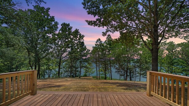 Une terrasse en bois, dotée de rampes de protection, qui mène à une zone herbeuse entourée d’arbres et offrant une vue sur une étendue d’eau.