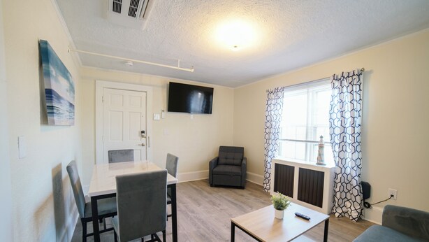 A modern living room with a dining table, two grey chairs, a grey sofa, a small coffee table, a grey armchair, a flat-screen TV mounted on the wall, and patterned curtains on the window. - Renovated One Bedroom Apartment Downtown Falmouth Minutes From the Beach  (Falmouth)