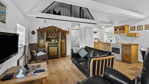 A spacious living area with a high ceiling, a dining table, a sofa, a television, a kitchen area with wooden cabinets, and a staircase leading to an upper level.