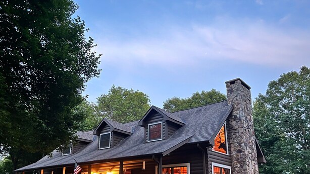 A log cabin with a stone chimney, multiple windows, and a porch with a railing. - Mountaintop at Yonahlossee Saddle (Boone)