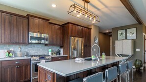 A modern kitchen with wooden cabinets, stainless steel appliances, a central island with bar stools, and a decorative light fixture. - Camp Cotter at Blue Ridge Mountain (Lenoir)
