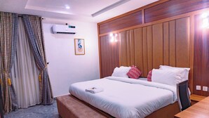 A neatly made bed with white linens and a small bedside table, a wooden headboard, and a wall-mounted air conditioner. - Rolarz Resorts & Hotel (Benin City)