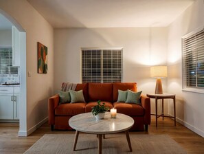 Una sala de estar con un sofá, una mesa redonda de centro, una lámpara y una ventana con persianas.