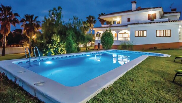 A swimming pool with a white deck, surrounded by grass and palm trees, with a white house in the background.