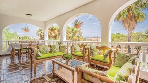A sunlit lounge area with wooden furniture, green cushions, and large arched windows offering views of palm trees and greenery.