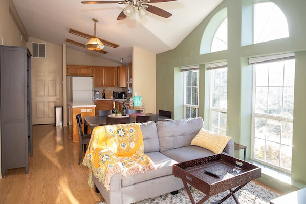 A spacious living area with a sofa, a coffee table, and a dining table. There is a kitchen in the background with wooden cabinets and a window.
