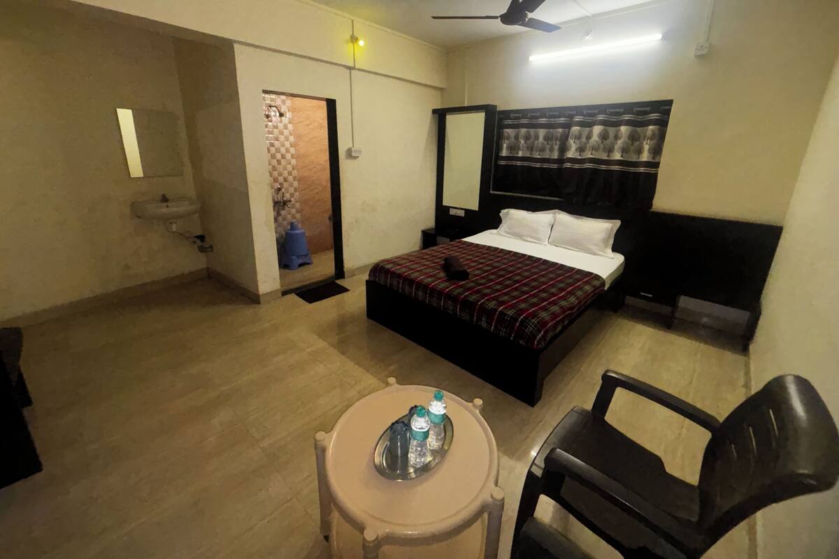 A hotel room with a bed, a chair, a small table with water bottles, a ceiling fan, and a bathroom with a sink and toilet.