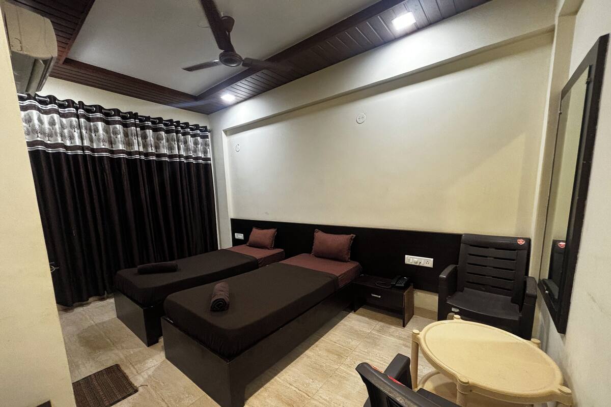 A hotel room with two beds, a small table, a chair, a ceiling fan, and a window with curtains.