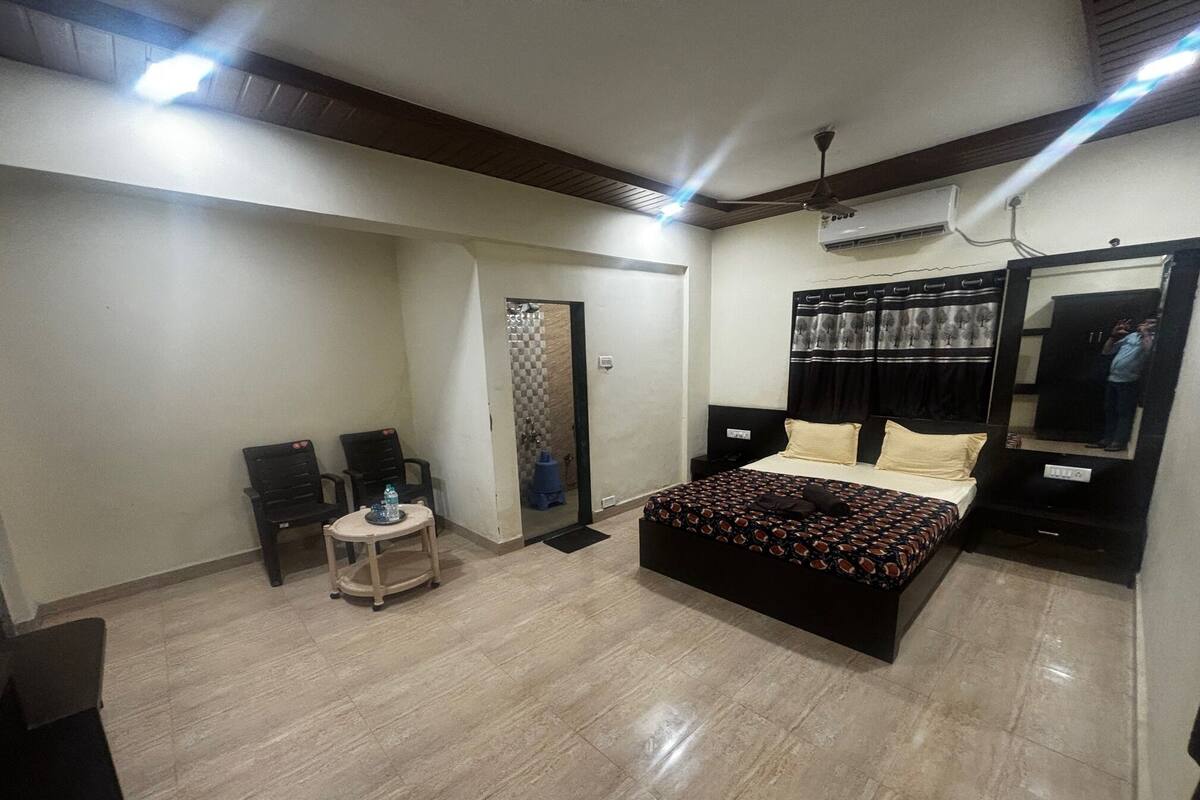 A bedroom with a bed, a small table with a water bottle, two chairs, a mirror, and an air conditioning unit.
