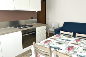 A compact kitchen with a gas stove, oven, and white cabinetry. There is a dining area with a table covered in a vintage car print tablecloth.