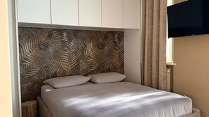 Ein Bett mit einem Kopfstück mit Blättermotiv, ein an der Wand montierter Fernseher und ein Fenster mit Vorhängen.