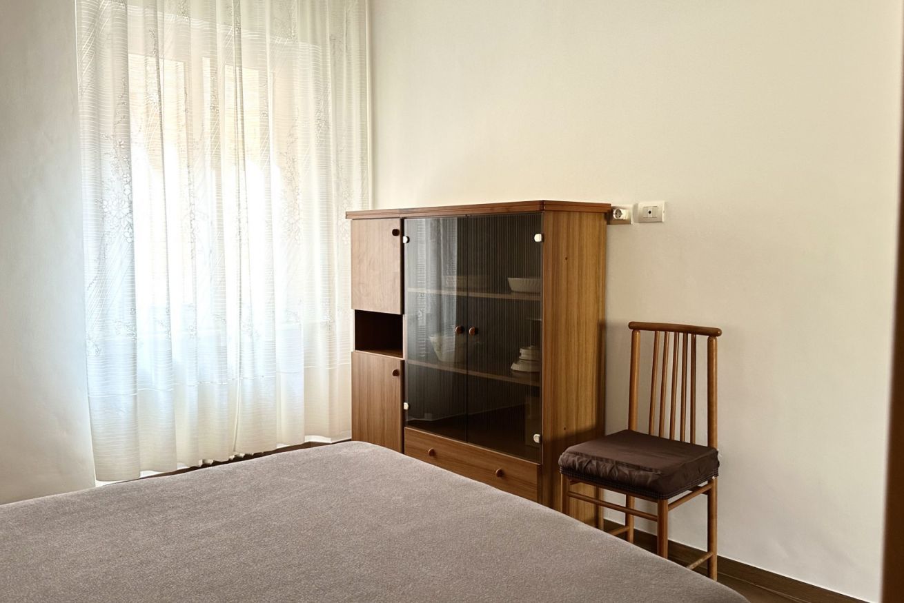 A bedroom with a bed, a wooden wardrobe with glass doors, a chair, and a window with sheer curtains.