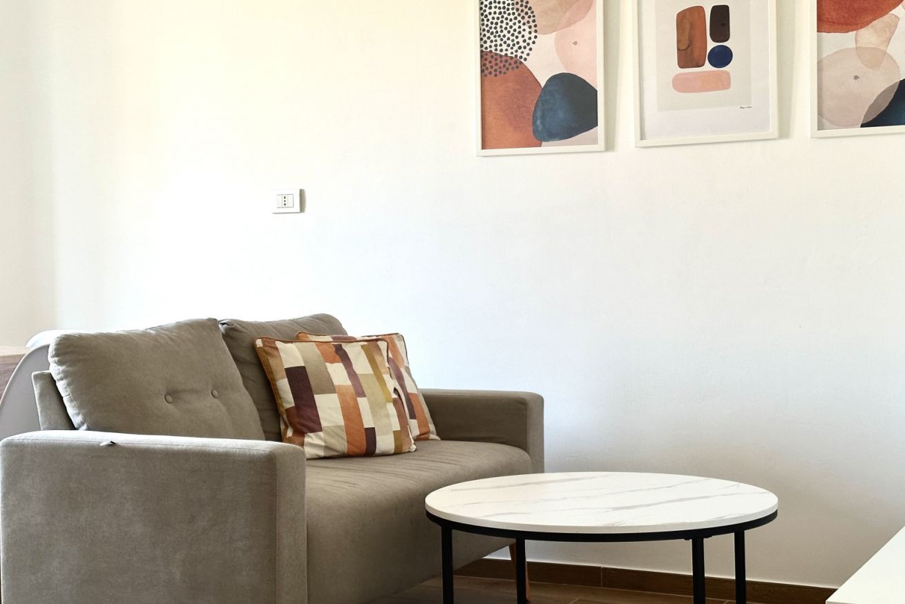 Ein Wohnzimmer mit grauem Sofa, einem gemusterten Kissen, einem runden weißen Tisch und abstrakter Kunst an der Wand.