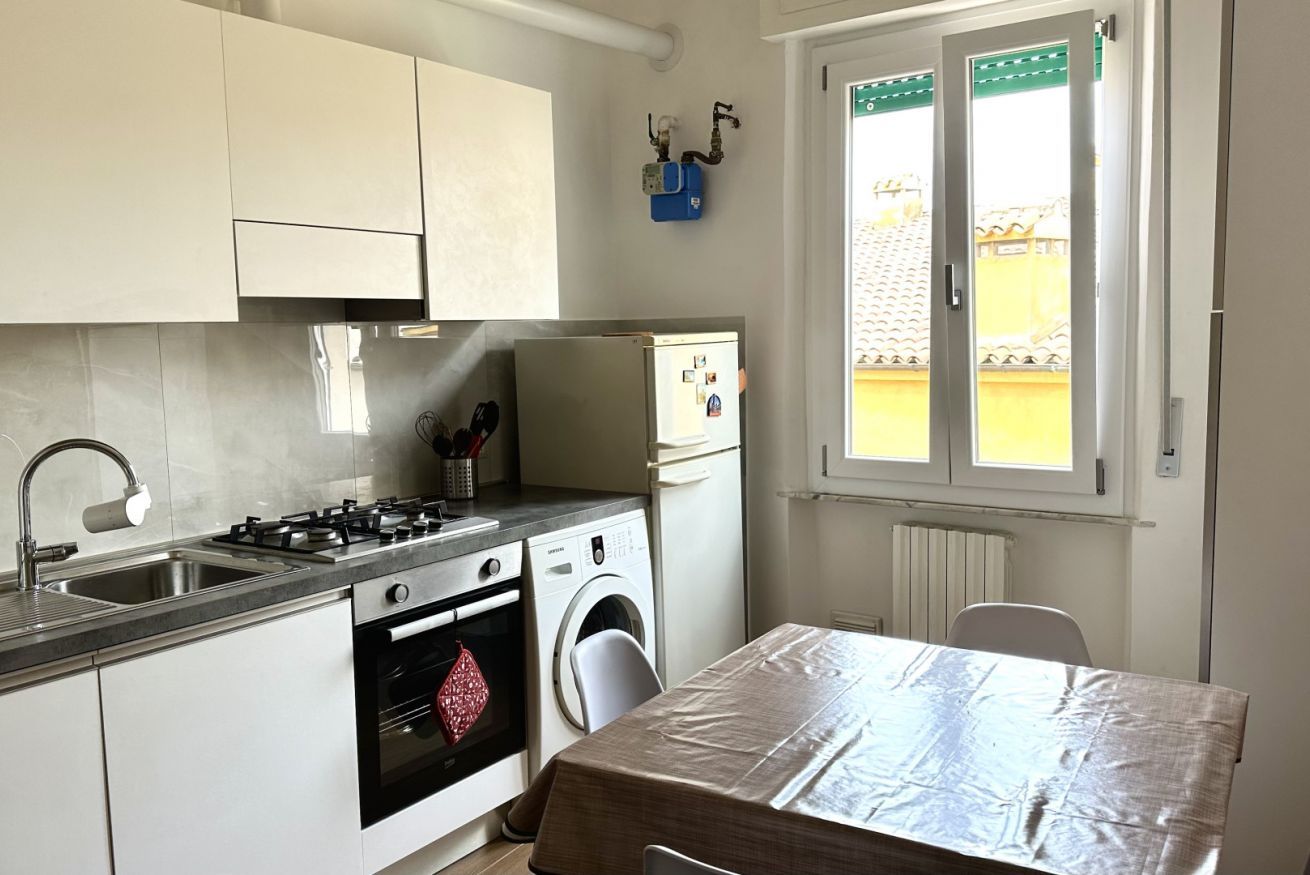 A compact kitchen with a gas stove, oven, washing machine, and a window with a view of a yellow building.
