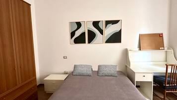 A bedroom with a bed, a desk, a chair, a wardrobe, and abstract wall art.
