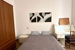 A bedroom with a bed, a desk, a chair, a wardrobe, and abstract wall art.