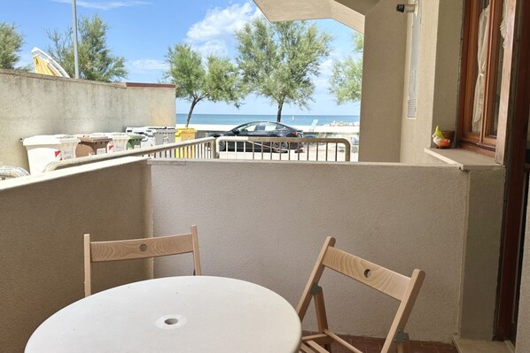 Un balcon avec une table ronde et deux chaises, donnant sur une plage et des voitures garées.