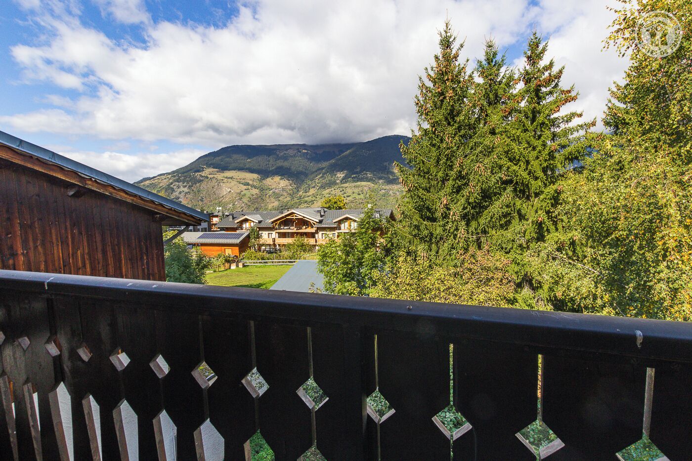 Un bâtiment en bois doté d’un balcon, un chemin menant à une rangée de maisons et une montagne en arrière-plan.