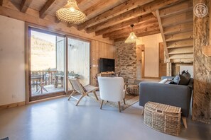 Een gezellige woonkamer met een stenen openhaard, houten balken in het plafond en een groot raam met uitzicht op een balkon.