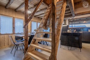 Een rustiek interieur met zichtbare houten balken, een eethoek met zwarte stoelen, een keuken met barstoelen, en grote ramen.