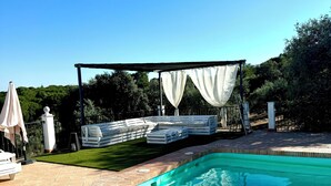 A swimming pool with a brick border, a covered seating area with white cushions, and a grassy lawn. - Casa Sierra Morena. Lince (AN)