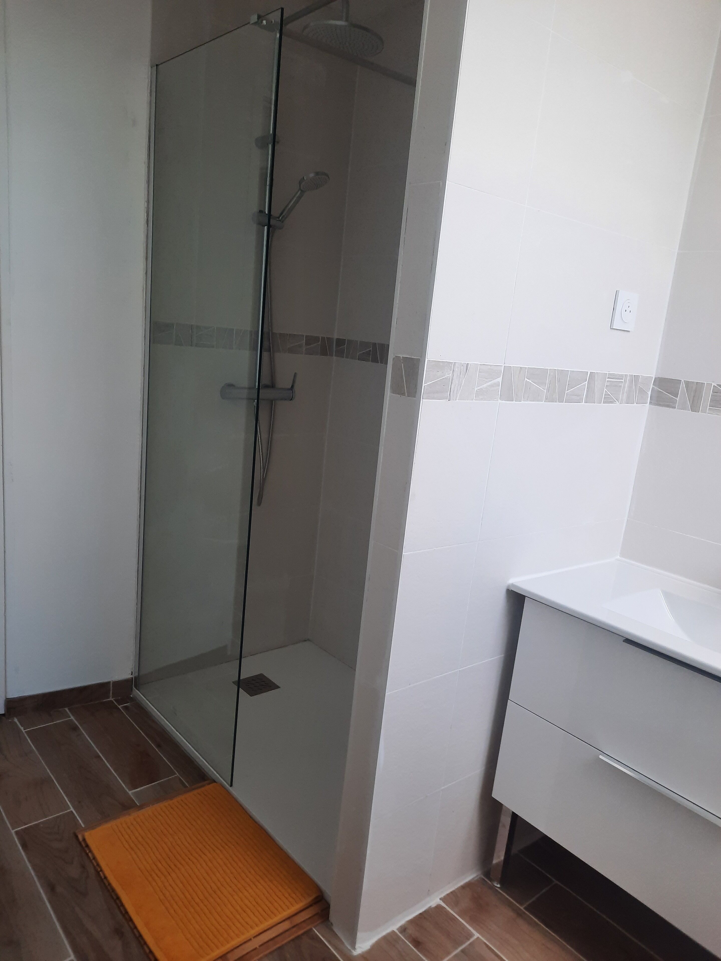 Ein Badezimmer mit einer gläsernen Dusche, einem weißen Waschbecken und einer Holzbadematte auf dem Boden.