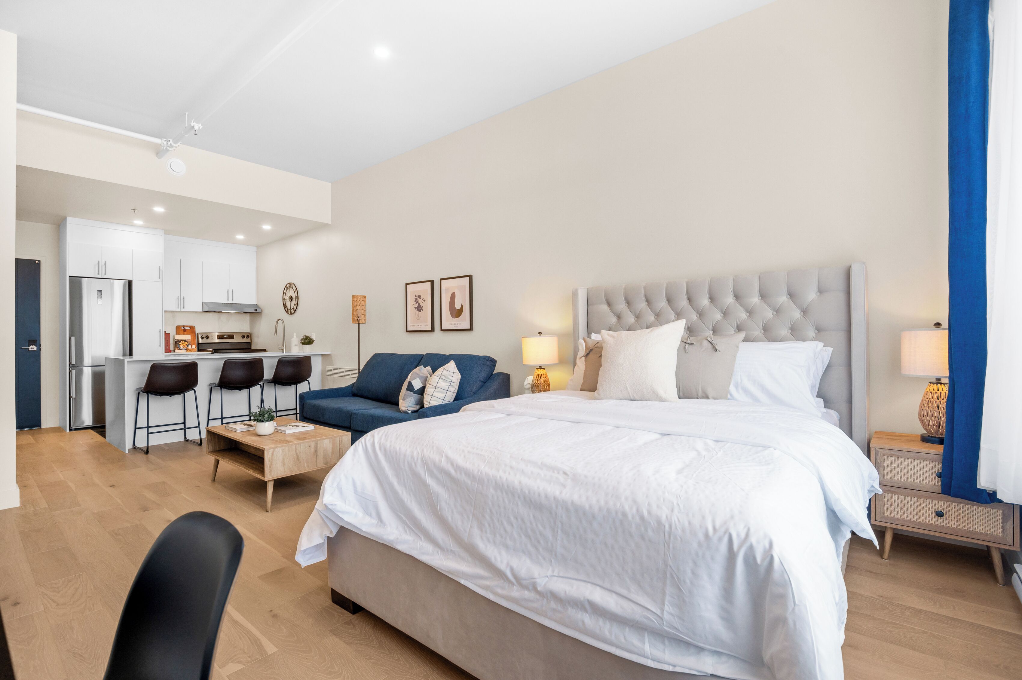 A modern hotel room with a large bed, a sofa, a dining area, and a kitchenette.