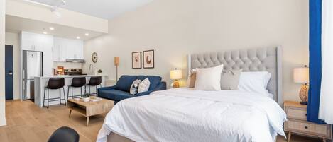 A modern hotel room with a large bed, a sofa, a dining area, and a kitchenette.