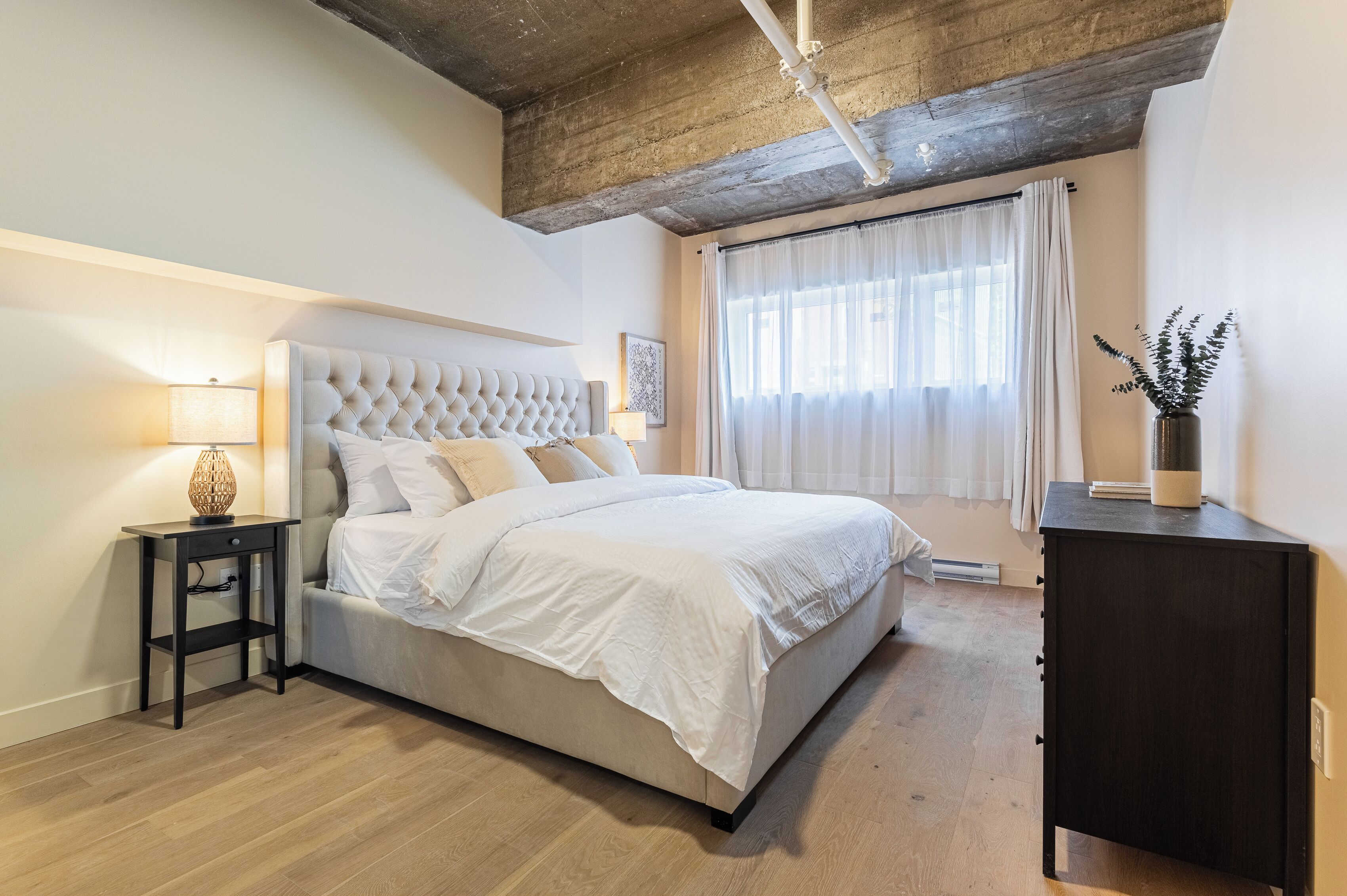 Een slaapkamer met een bed met een gestuwd hoofdeinde, een nachtkastje met een lamp, een donker houten dressoir en een groot raam met dunne gordijnen.