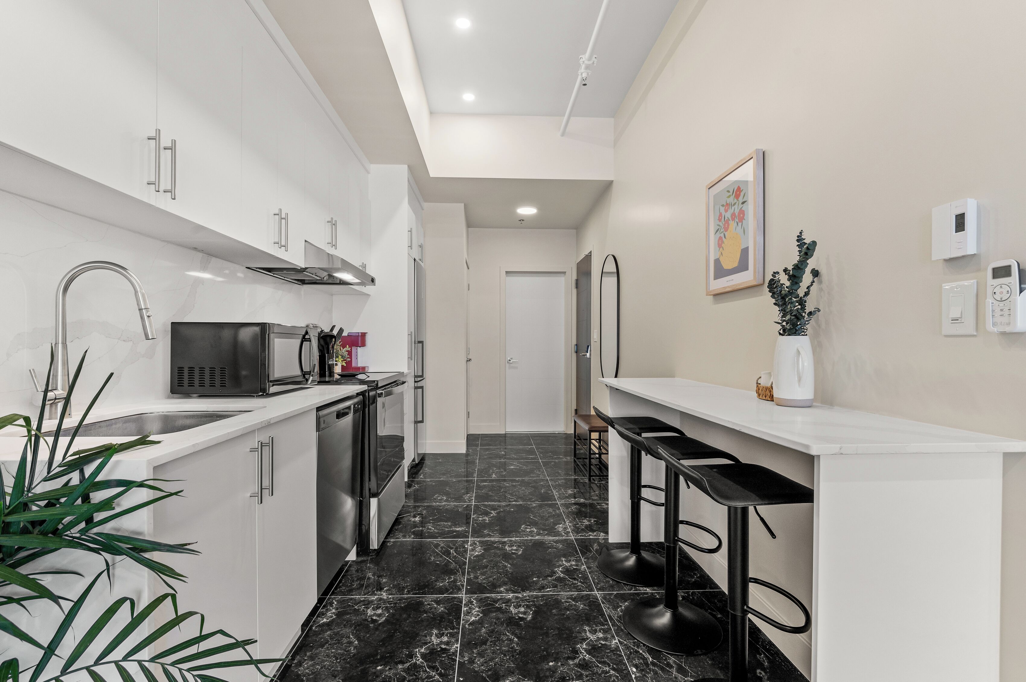 A modern kitchen with white cabinets, a black countertop, and a black tiled floor. There are two black bar stools and a white countertop with a plant.