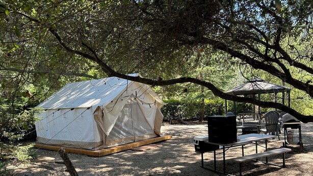 Una carpa montada en un área boscosa, con una mesa de picnic y una parrilla cerca.