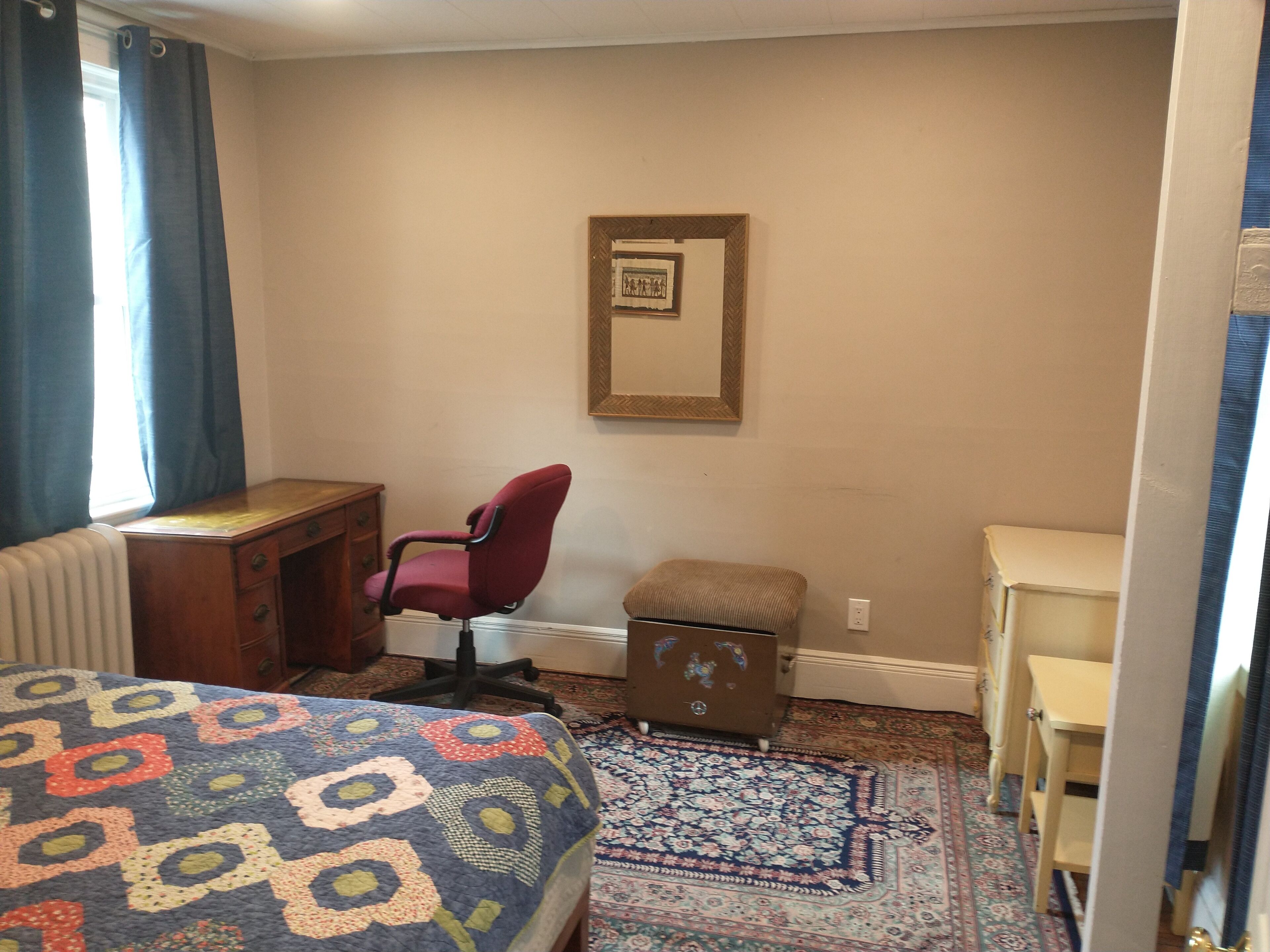 A hotel room with a bed, a desk, a chair, a radiator, a framed picture on the wall, and a small table.