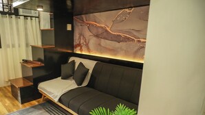 A modern bedroom with a dark sofa, a small bedside table, a wall-mounted shelf, and a large abstract painting.