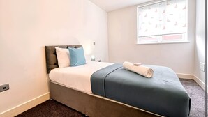 Un lit bien fait, avec un matelas gris et une literie bleue, une table de chevet avec une lampe, et une fenêtre ornée d’un motif représentant un voilier.