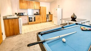 Une table de billard avec un queue de billard et des billes, un coin cuisine avec des meubles en bois et des appareils électroménagers, et une petite table à manger agrémentée d’un vase.