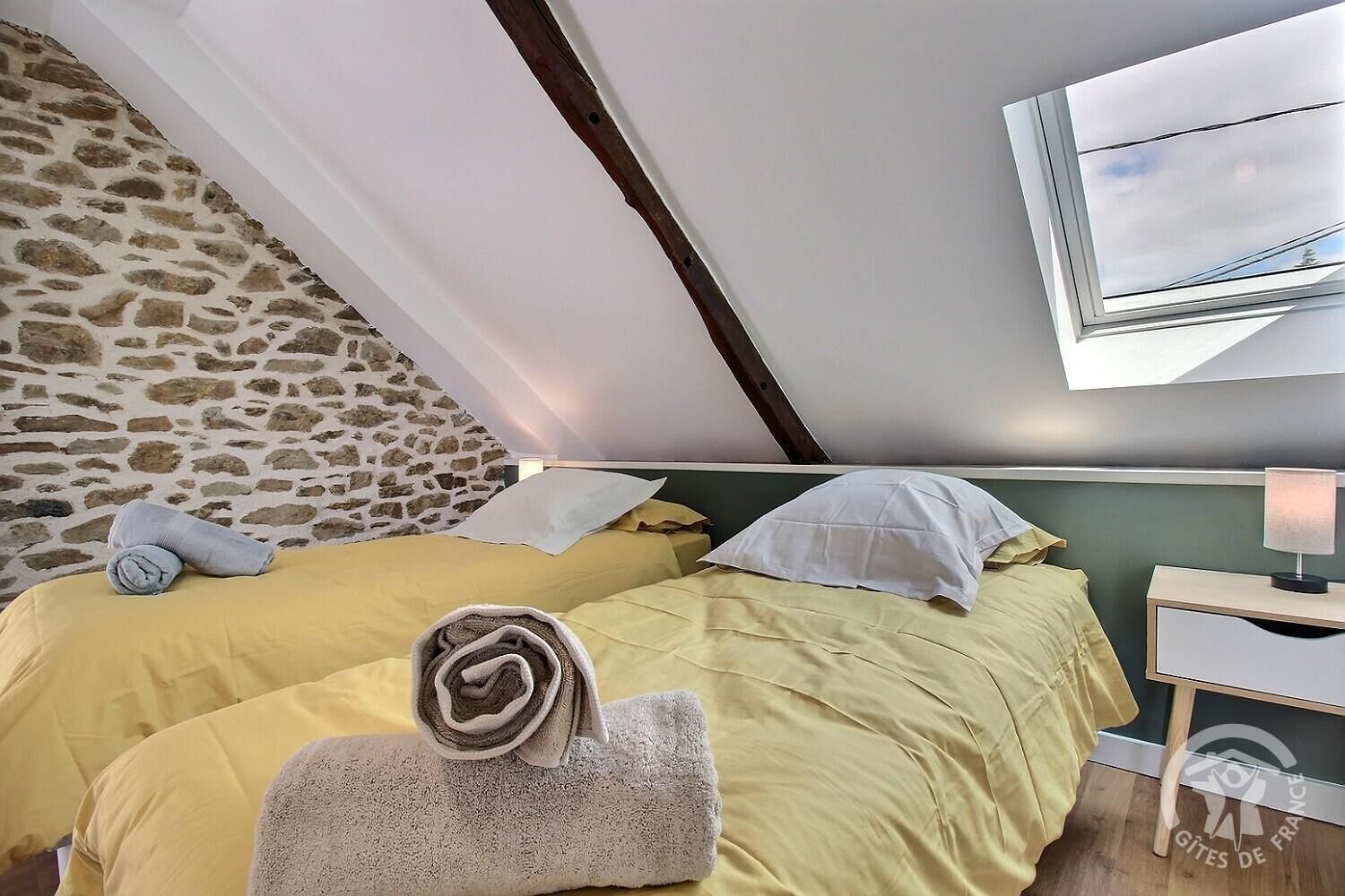 A bedroom with two beds, a stone wall, a skylight, and a bedside table with a lamp.
