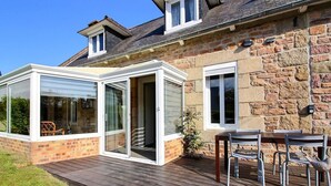 Una casa con porche acristalado, fachada de piedra y terraza de madera con mobiliario exterior.