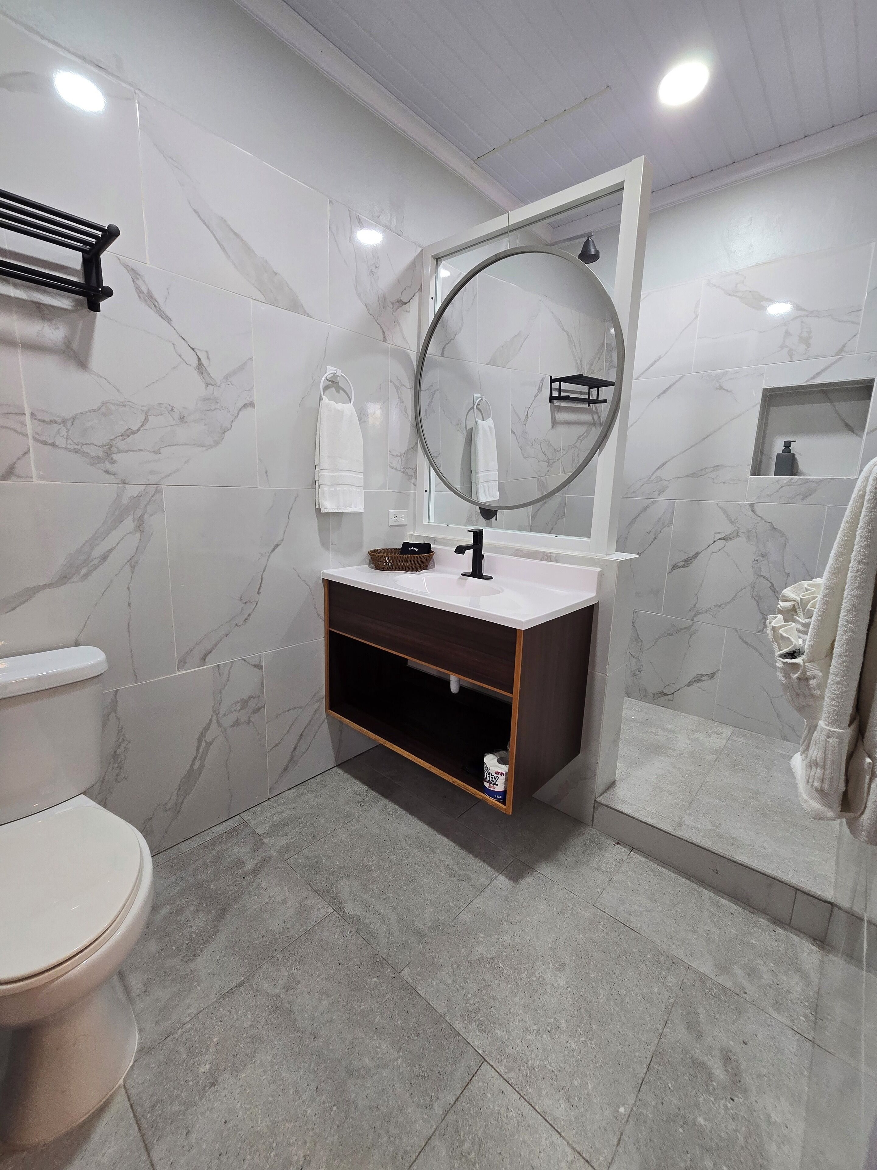 Een moderne badkamer met een betegelde muur van marmer, een ronde spiegel, een witte wasbak en een toilet.