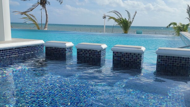 A pool with clear blue water and tiled edges, surrounded by palm trees and a view of the ocean. - Island Magic Villas (Caye Caulker)