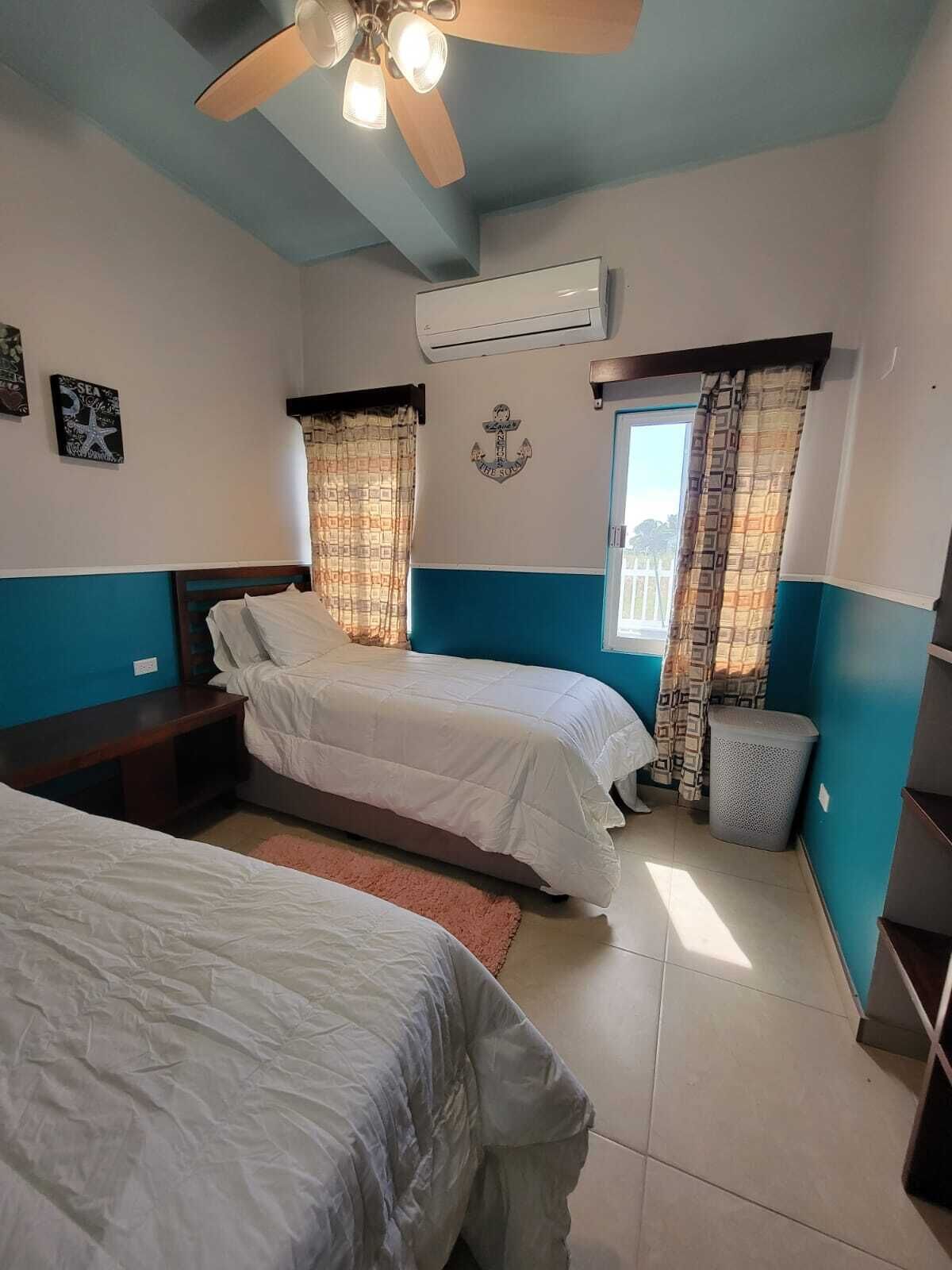 Habitación de hotel con dos camas, ventilador de techo, equipo de aire acondicionado y ventana con cortinas.