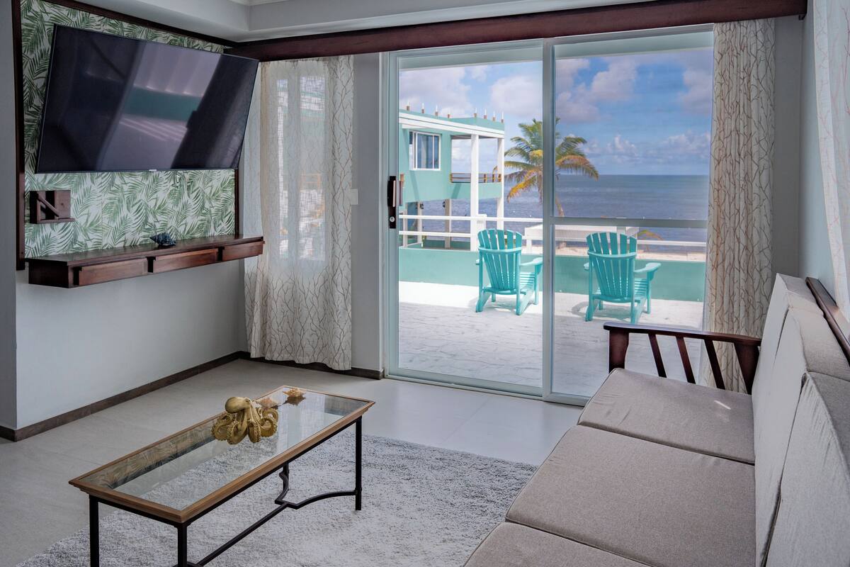 Ein Wohnzimmer mit Fernseher, einem Glastisch und einer Couch. Eine Glastür führt auf einen Balkon mit Sitzgelegenheiten und Blick auf das Meer.