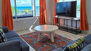 A modern living room with a large TV, two sofas, a round coffee table, and a view of the ocean through large windows with orange curtains. - Island Magic Villas (Caye Caulker)