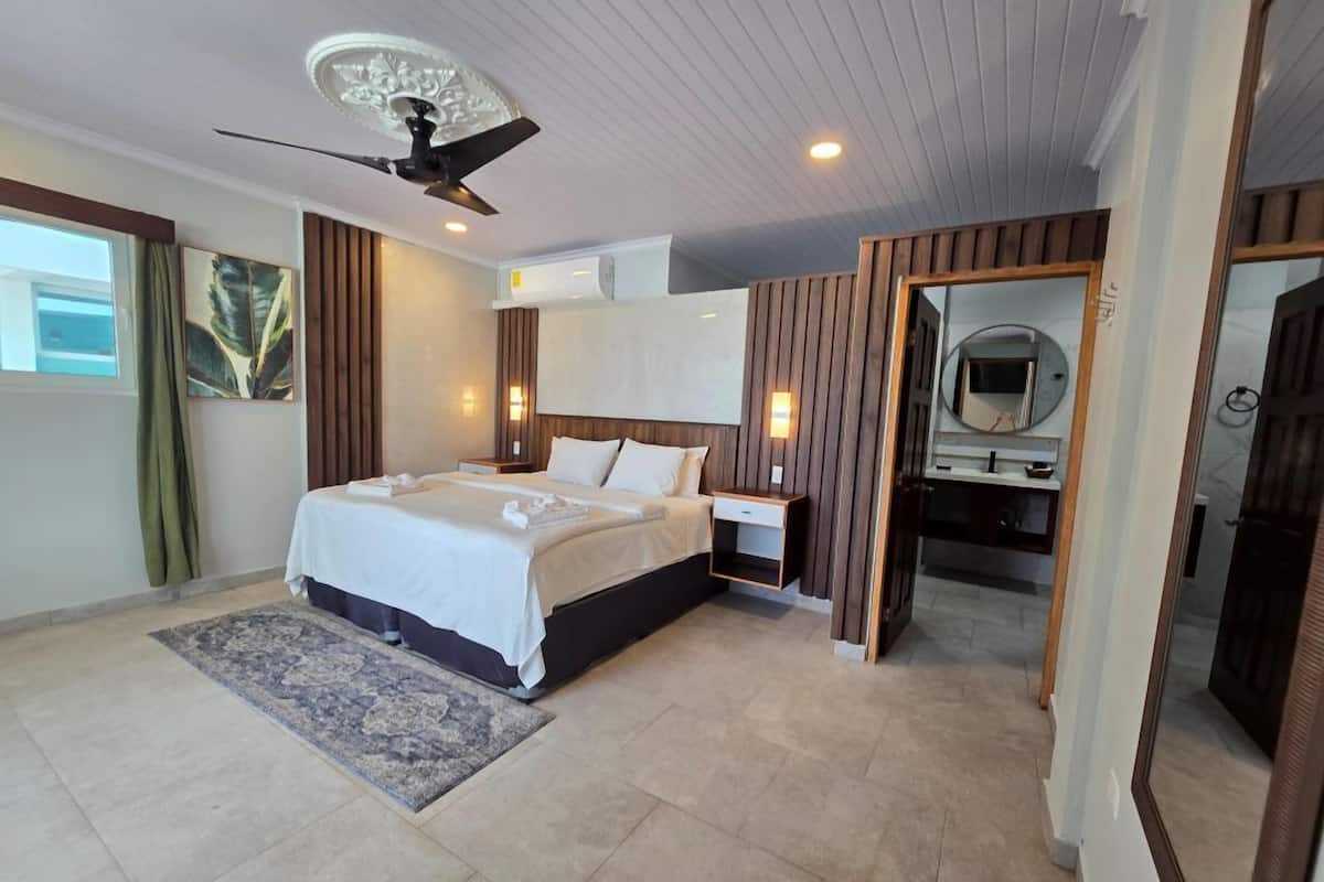 Ein modernes Hotelzimmer mit einem großen Bett, einem Deckenventilator, einer Wand-Klimaanlage und einem Badezimmer mit Spiegel und Toilette.