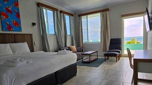 A hotel room with a large bed, a sofa, a dining table, and a chair. There are windows with curtains, a painting on the wall, and a view of the sea. - Island Magic Villas (Caye Caulker)